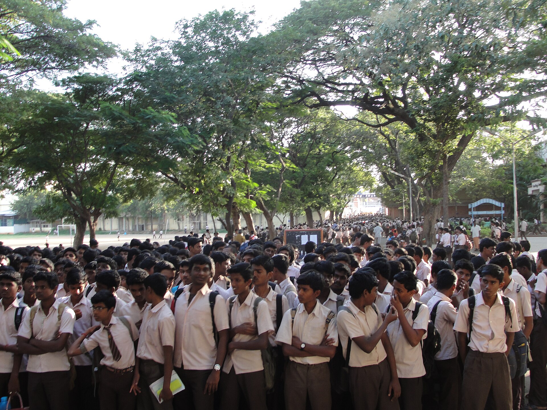 High-school students in white shirts gathered on a tree-lined school campus in India