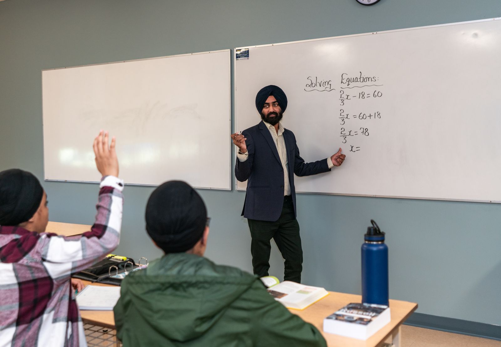 Teacher solving equations on a whiteboard while a student raises a hand