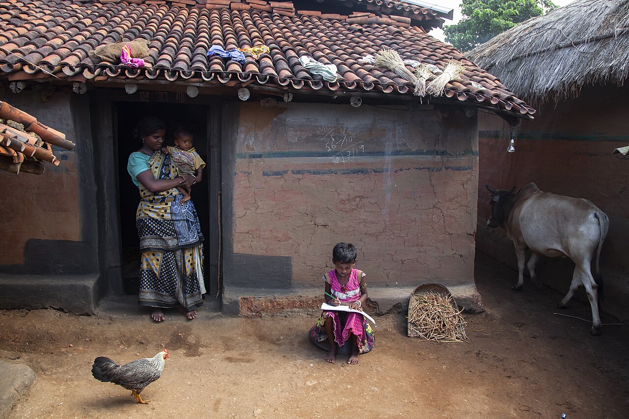 A girl studying outside her home while her mother stands at the door holding a baby