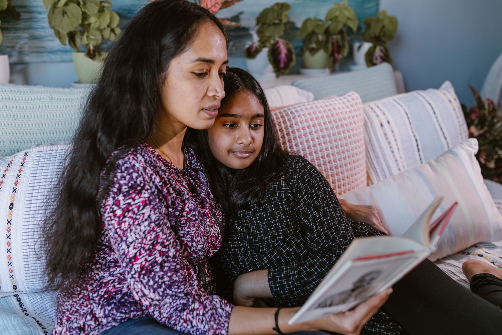 Indian mother and teenage daughter reading a book together on a sofa at home