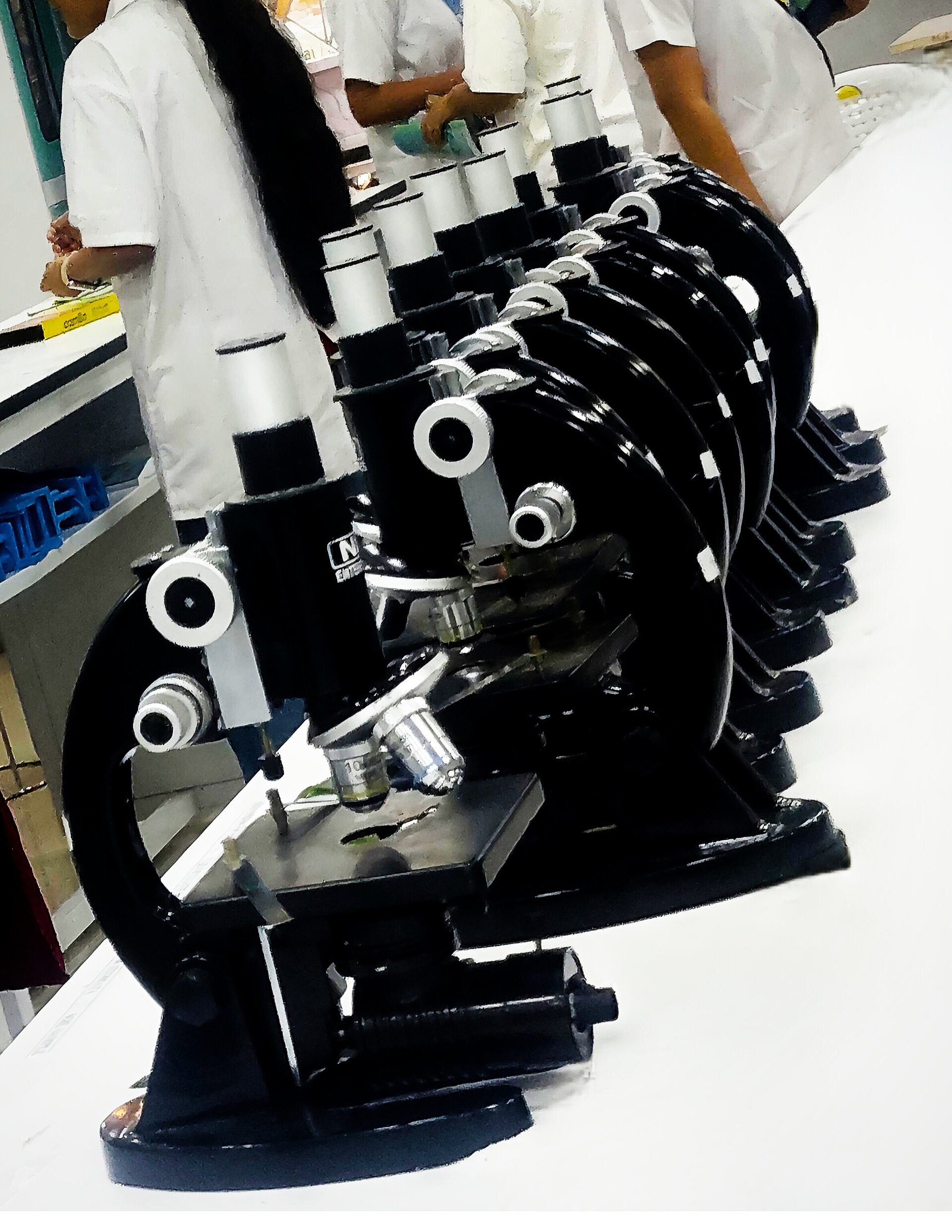 A row of compound microscopes on a laboratory bench with students in white coats in the background