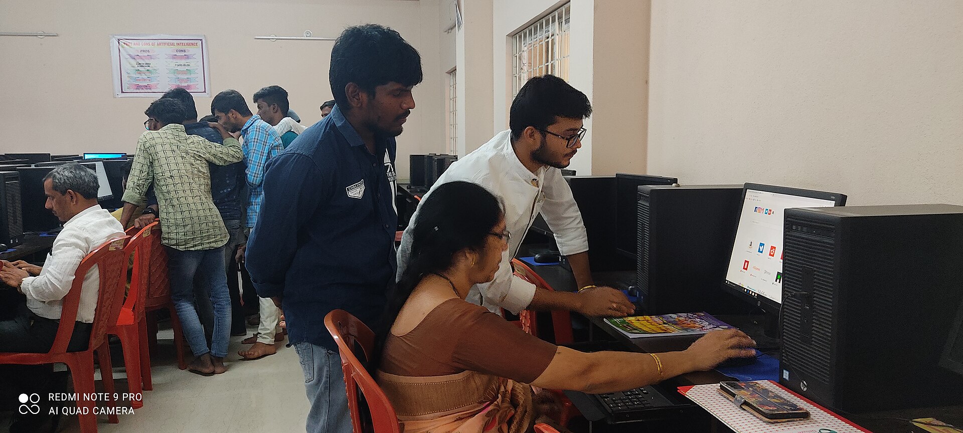 A woman uses a desktop computer while a young man guides her in a college computer lab