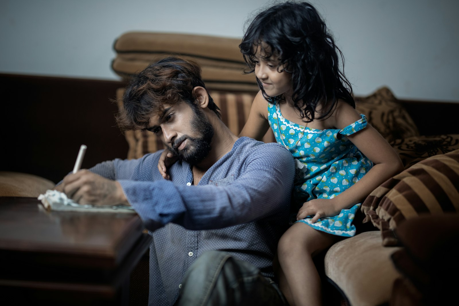 A father works through a notebook at home while his daughter watches closely.